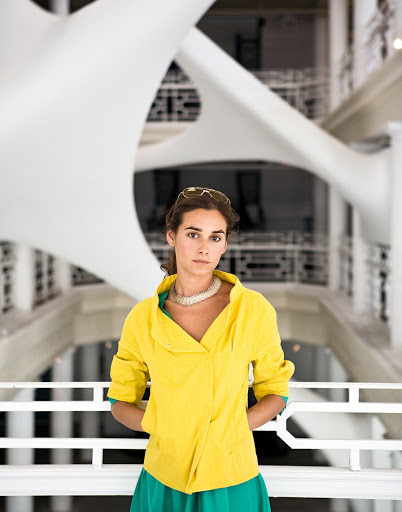 Indoor portrait of a woman in a yellow jacket and green skirt, wearing a white necklace, with a white sculpture in the background.