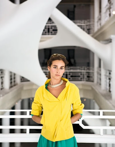 Indoor portrait of a woman in a yellow jacket and green skirt, wearing a white necklace, with a white sculpture in the background.