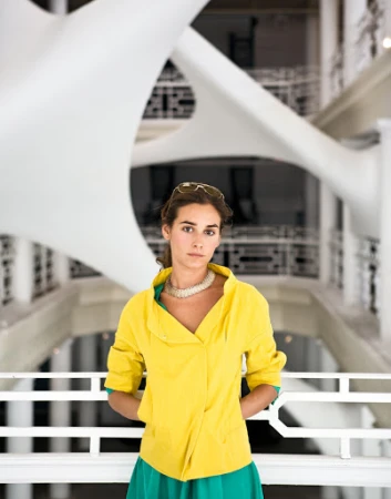 Indoor portrait of a woman in a yellow jacket and green skirt, wearing a white necklace, with a white sculpture in the background.