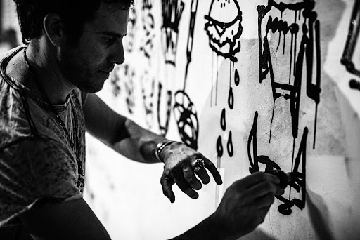 Adult male artist creating black-and-white graffiti-style painting on a white wall with a marker indoors, monochrome.