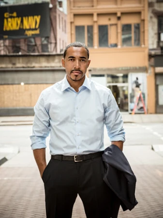 Portrait of a man in a button-down shirt and dark slacks standing in a city street with a building facade in the background.
