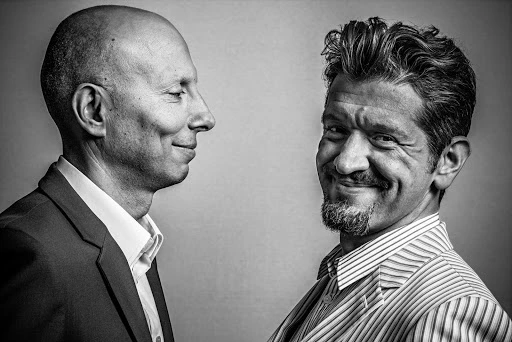 Black and white portrait of two adult men indoors. One man is bald and wearing a suit, while the other has a beard and a striped shirt.