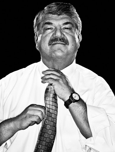 Black and white portrait of an adult male adjusting his patterned necktie, wearing a white shirt and a watch on a black background.
