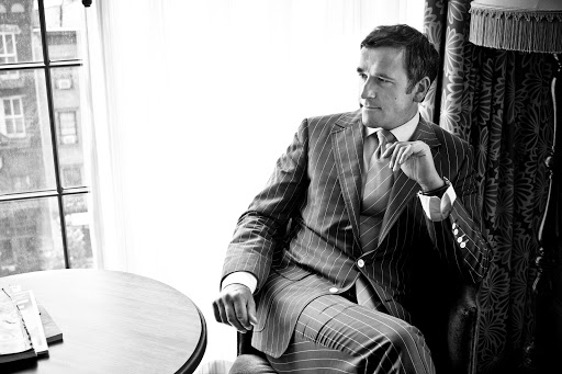 Black and white portrait of a man in a striped suit sitting indoors near a window and round table, exuding classic elegance.