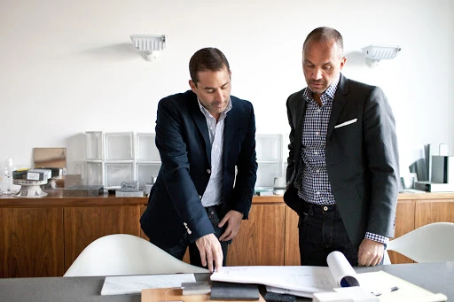 Two adult men in business suits review documents at a table in a bright office. Architectural plans and material samples visible.