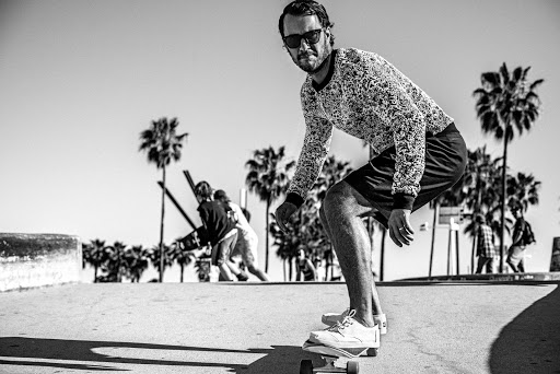 Man skateboarding on a sunny day in an outdoor park setting with palm trees in the background. Wearing a patterned shirt and shorts.