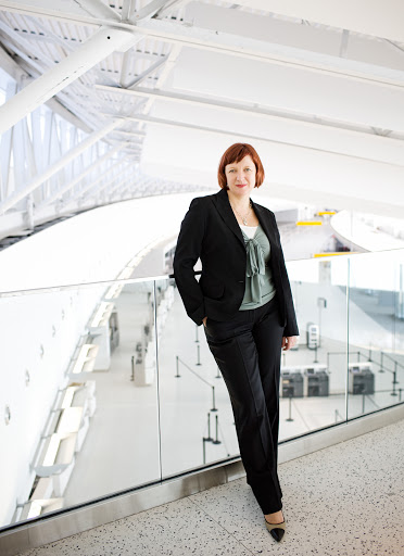 Professional woman with auburn hair poses in a black suit against a modern white airport backdrop filled with natural light.