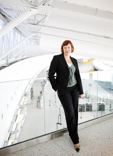 Professional woman with auburn hair poses in a black suit against a modern white airport backdrop filled with natural light.