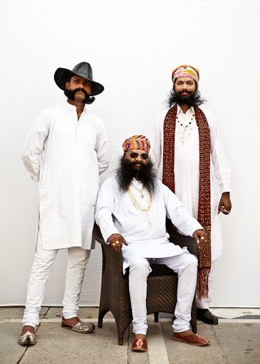 Three bearded men in traditional white clothing. One man sits in a chair, wearing a turban and sunglasses, while the other two stand behind.