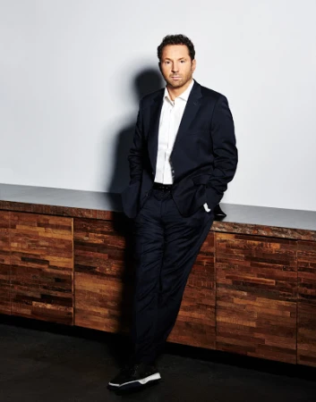 Man in dark suit and white dress shirt leaning against a wood credenza in a studio