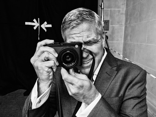 Monochrome portrait of George Clooney holding a Leica camera with focus on the camera lens, in a photo studio setting, looking at the camera.