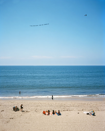 Beach scene with people sunbathing on sand, ocean waves, sky with banner plane. Summertime coastal view of relaxation by the sea.