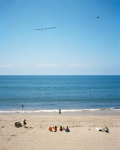 Beach scene with people sunbathing on sand, ocean waves, sky with banner plane. Summertime coastal view of relaxation by the sea.