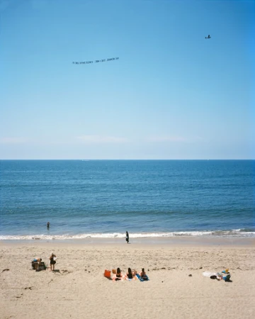 Beach scene with people sunbathing on sand, ocean waves, sky with banner plane. Summertime coastal view of relaxation by the sea.