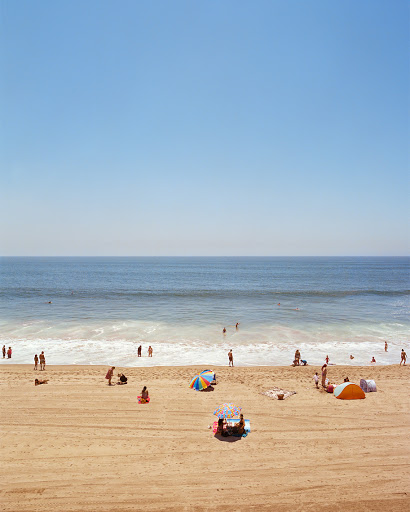 Beach scene with numerous people relaxing and enjoying the sunny weather near the ocean under a clear sky.