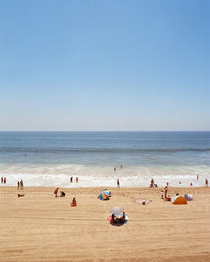Beach scene with numerous people relaxing and enjoying the sunny weather near the ocean under a clear sky.