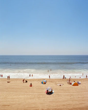 Beach scene with numerous people relaxing and enjoying the sunny weather near the ocean under a clear sky.
