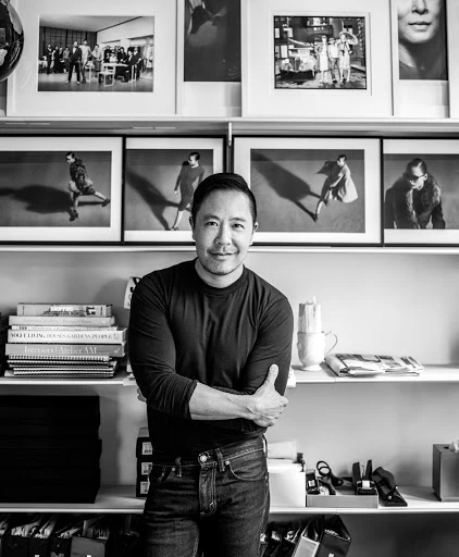 Black and white portrait of a smiling Asian man indoors in front of a shelving unit with framed artwork and stacked books.