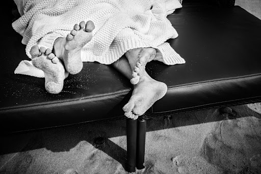 Black and white photograph of feet covered in sand protruding from under a blanket on a lounge chair on a sandy surface outdoors.