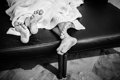 Black and white photograph of feet covered in sand protruding from under a blanket on a lounge chair on a sandy surface outdoors.