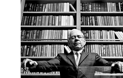 Black and white portrait of an adult male with short white hair and glasses, wearing a suit and tie, posed in front of a bookshelf.