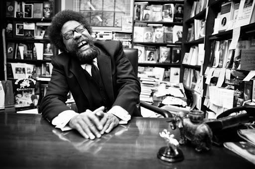 Portrait of adult male with glasses, beard, and afro laughing at a wooden desk with books in the background in black and white.
