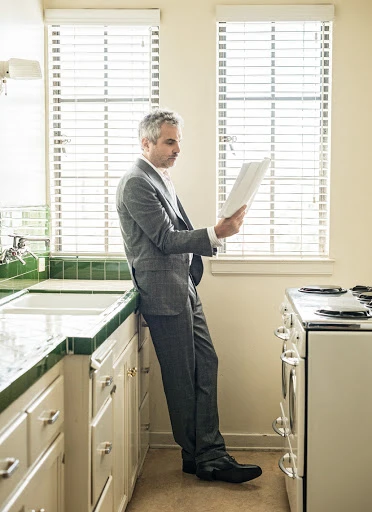 Adult male in a gray suit leans against a kitchen counter, reading documents by a window with blinds, near a stove and cabinets.