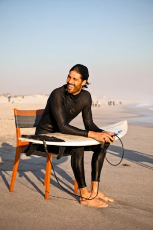 Smiling surfer wearing a black wetsuit seated on an orange chair on a sandy beach holding a surfboard
