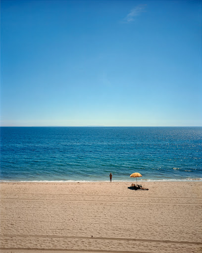 Beach scene featuring an ocean view, a sandy beach, blue skies, a person standing near the shoreline, and a beach umbrella.