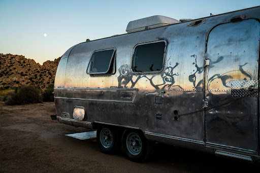 Vintage Airstream trailer parked in a desert landscape at dusk, reflecting the surrounding environment on its polished aluminum exterior.