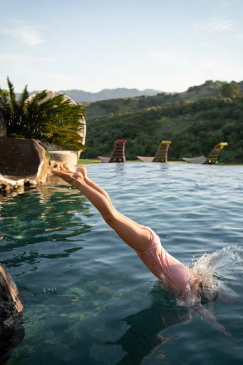 Adult female in a pink swimsuit dives into an outdoor pool with a tropical background and mountains on a clear, sunny day.