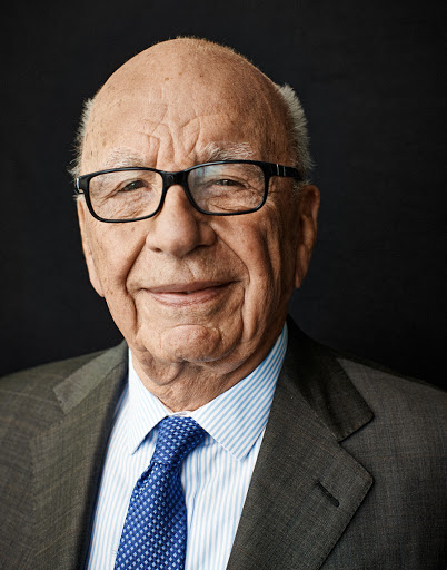 Headshot of an elderly adult male with glasses and a suit and tie against a dark grey backdrop in an indoor setting.