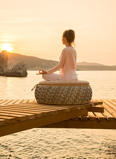 Adult female in lotus position meditating on woven cushion on wooden dock, calm waters, and scenic rock formations at sunset.