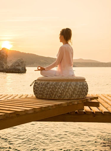 Adult female in lotus position meditating on woven cushion on wooden dock, calm waters, and scenic rock formations at sunset.