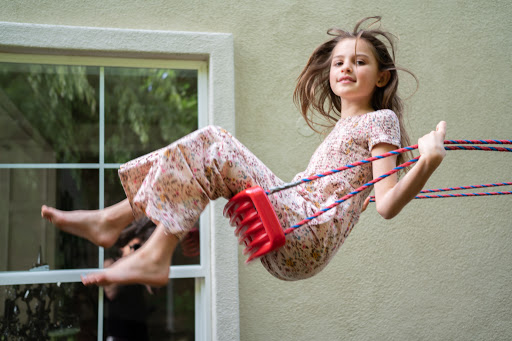 Girl on a swing outside, wearing floral print outfit, with a window and green wall in background, creating a playful outdoor scene.