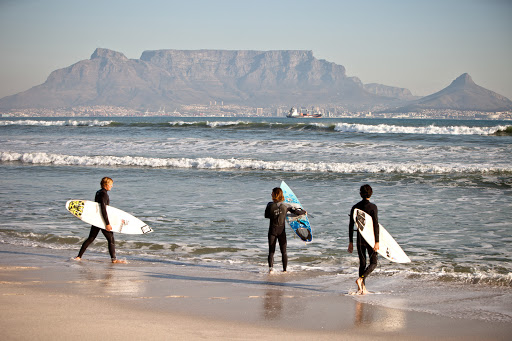 Three young surfers in wetsuits walk on a beach with surfboards, ocean waves, and a mountain range in the background on a sunny day.