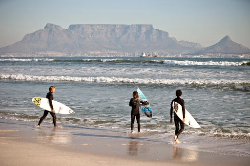 Three young surfers in wetsuits walk on a beach with surfboards, ocean waves, and a mountain range in the background on a sunny day.