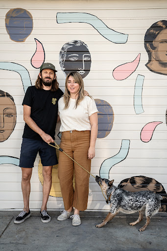 A stylish couple and their dog pose in front of a mural featuring abstract faces and shapes painted in neutral colors on a white wall.