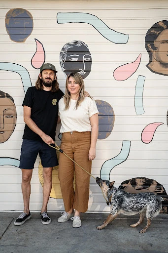 A stylish couple and their dog pose in front of a mural featuring abstract faces and shapes painted in neutral colors on a white wall.