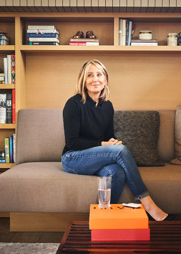 A woman with blonde hair sits on a sofa in front of a bookcase. She is wearing jeans and a dark top. A glass of water sits on a table.