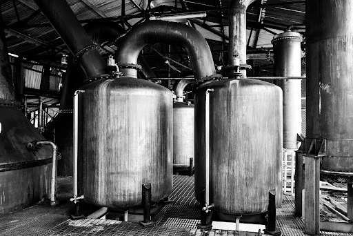 Monochrome photograph of metal industrial tanks, pipes, and framework inside a distillery, showcasing the machinery used in spirit production.