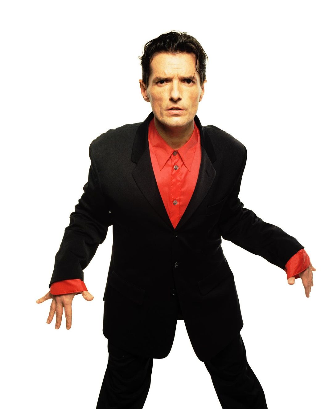Studio shot of a man in a black suit and red shirt, standing against a plain white backdrop with an outstretched arm pose.