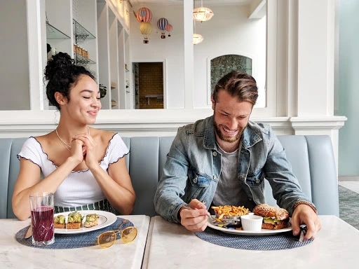 A young couple sits in a restaurant, a woman with curly hair smiles at the man as he eats a burger and fries at a table.
