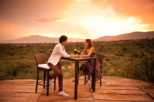 Couple enjoys drinks at a table on a wooden deck, with a lush green landscape and a vivid sunset in the background.