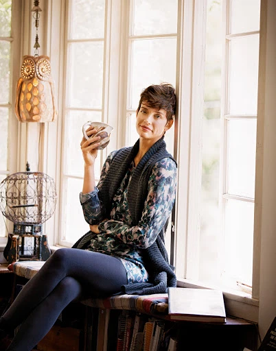 Woman with short hair sits in front of a large window, holding a mug. Natural light floods the interior space, showcasing the casual setting.