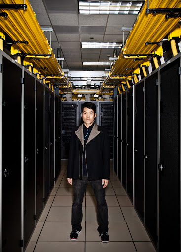 Man stands in a server room with black server racks lining the walls, bright yellow cables running overhead in an indoor setting.