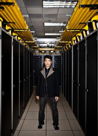 Man stands in a server room with black server racks lining the walls, bright yellow cables running overhead in an indoor setting.