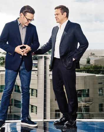 Two men in business attire standing on a rooftop terrace overlooking an urban skyline under daylight.