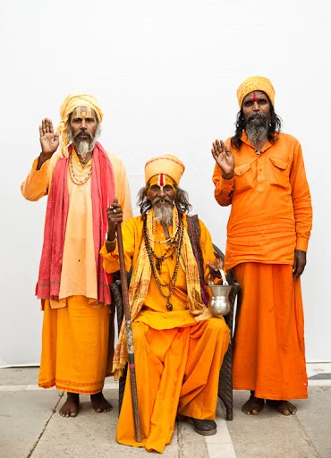 Three Indian men in traditional orange robes pose outdoors against a bright white background, one seated and holding a silver pot.