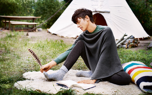 Young man in outdoor setting with tent, seated on a fur rug, wearing a poncho, reading a book, holding a feather in his hand.